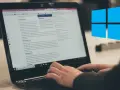 Ręce piszą na klawiaturze laptopa, na ekranie widać artykuł o malowaniu dachu. Obok logo Windows.