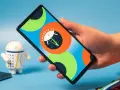 Dłoń trzyma smartfon z logo Android 14. Obok stoi figurka robota.