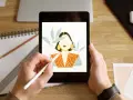 Ręce trzymają tablet, na którym wyświetla się portret kobiety w stylu procreate android. Obok leży notatnik i ołówek.