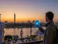Mężczyzna z plecakiem trzyma telefon z napisem "5G CONNECT" na tle zachodzącego słońca nad Nilem, z widocznymi piramidami w Egipcie. eSIM Egipt zapewnia łączność.