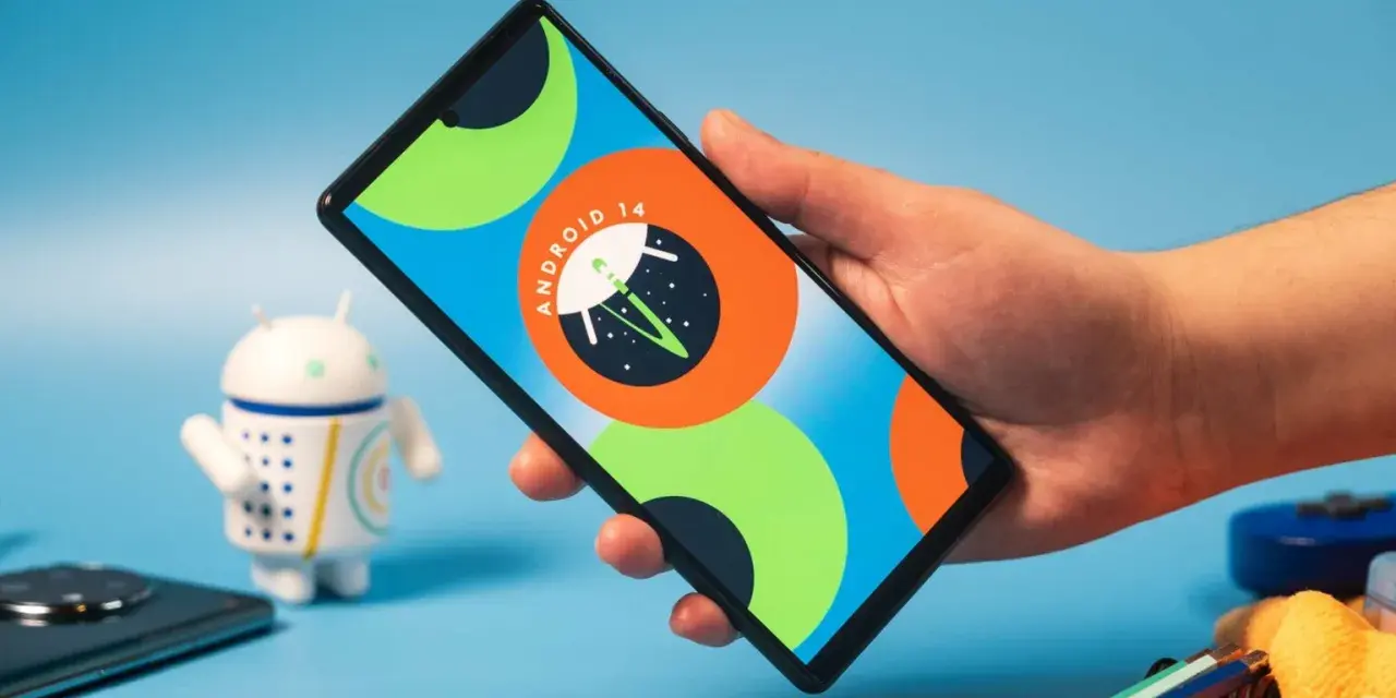 Dłoń trzyma smartfon z logo Android 14. Obok stoi figurka robota.