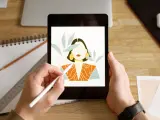 Ręce trzymają tablet z rysunkiem kobiety, tworzonym w aplikacji procreate. Obok leżą szkicownik i ołówek.