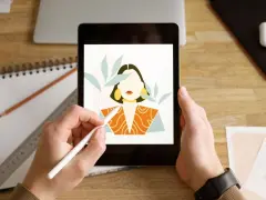 Ręce trzymają tablet z rysunkiem kobiety, tworzonym w aplikacji procreate. Obok leżą szkicownik i ołówek.