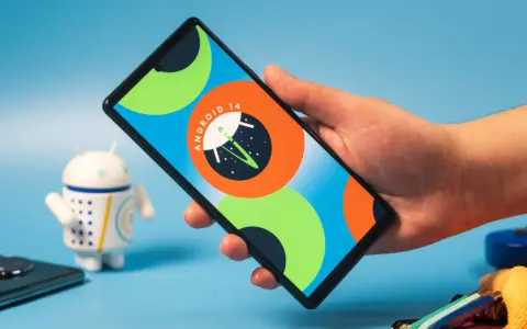 Dłoń trzyma smartfon z logo Android 14. Obok stoi figurka robota.