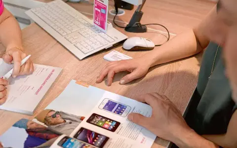Dwie osoby przeglądają ofertę telefonów komórkowych. Na biurku leży klawiatura, myszka, skaner i katalog z produktami. Jedna z osób pisze coś na formularzu.
