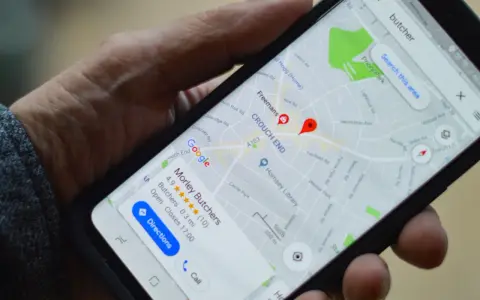 Dłoń trzyma smartfon z mapą Google. Widoczne są google maps współrzędne i wyszukiwanie "butcher".