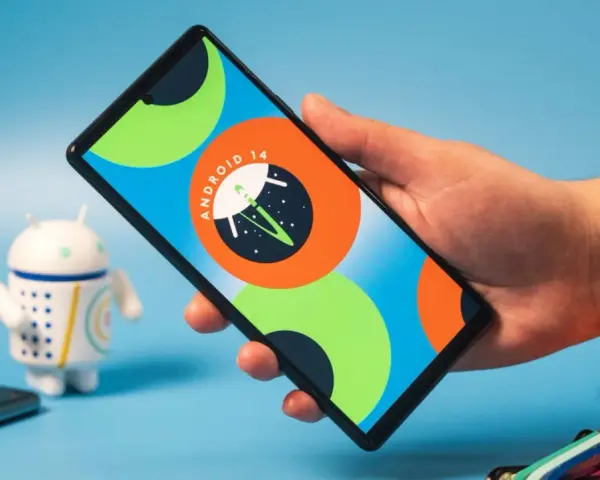 Dłoń trzyma smartfon z logo Android 14. Obok stoi figurka robota.
