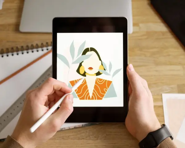 Ręce trzymają tablet, na którym wyświetla się portret kobiety w stylu procreate android. Obok leży notatnik i ołówek.