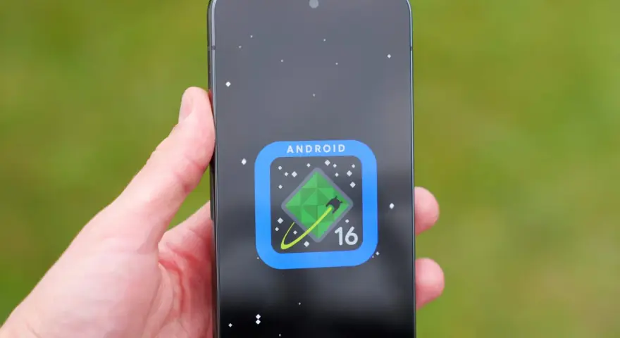 Dłoń trzyma smartfon z ikoną Android 16 na ekranie, przypominającą zielony kryształ na tle kosmosu.