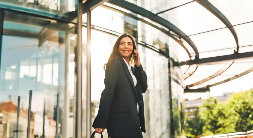 Uśmiechnięta kobieta w eleganckim stroju rozmawia przez telefon, niosąc torbę. Jej podróż po Wielkiej Brytanii trwa, a słońce odbija się w szklanej fasadzie.
