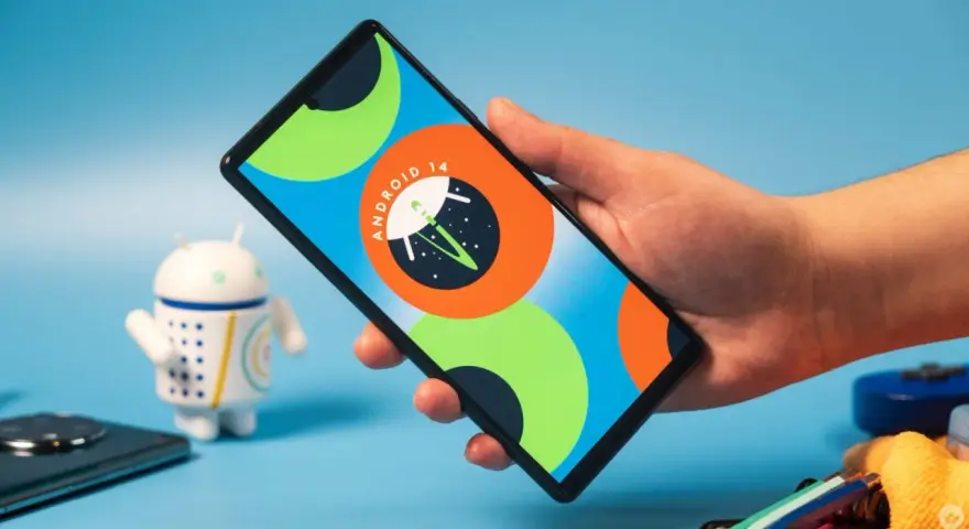 Dłoń trzyma smartfon z logo Android 14. Obok stoi figurka robota.