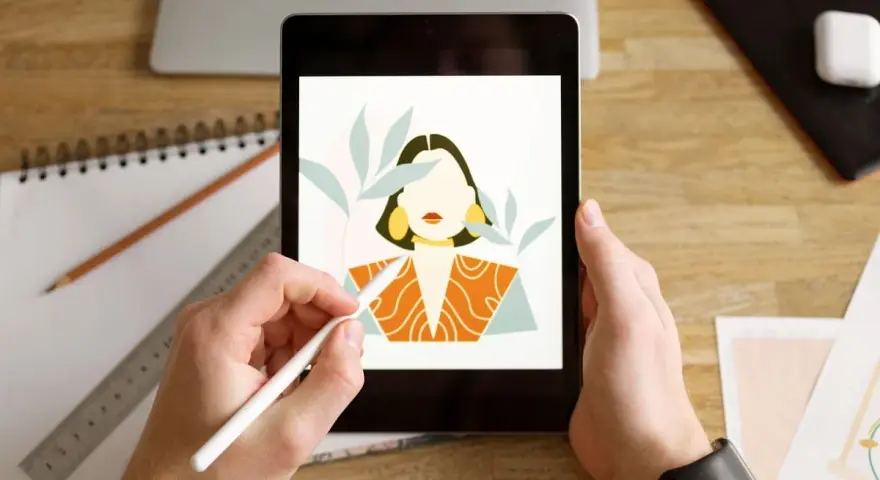 Ręce trzymają tablet, na którym wyświetla się portret kobiety w stylu procreate android. Obok leży notatnik i ołówek.