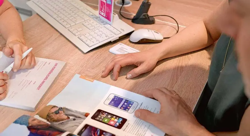Dwie osoby przeglądają ofertę telefonów komórkowych. Na biurku leży klawiatura, myszka, skaner i katalog z produktami. Jedna z osób pisze coś na formularzu.