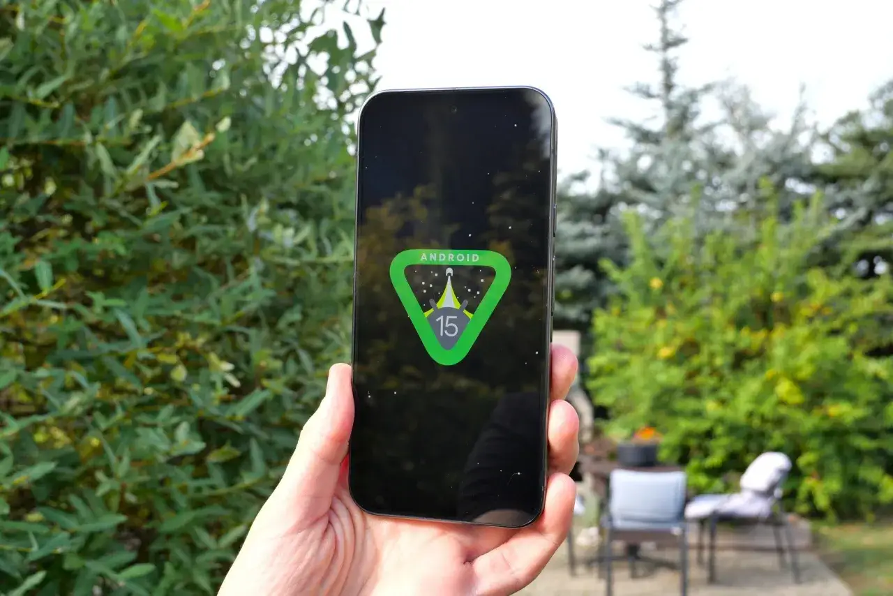 Dłoń trzyma smartfon z logo Android 15 na ekranie, na tle zieleni.