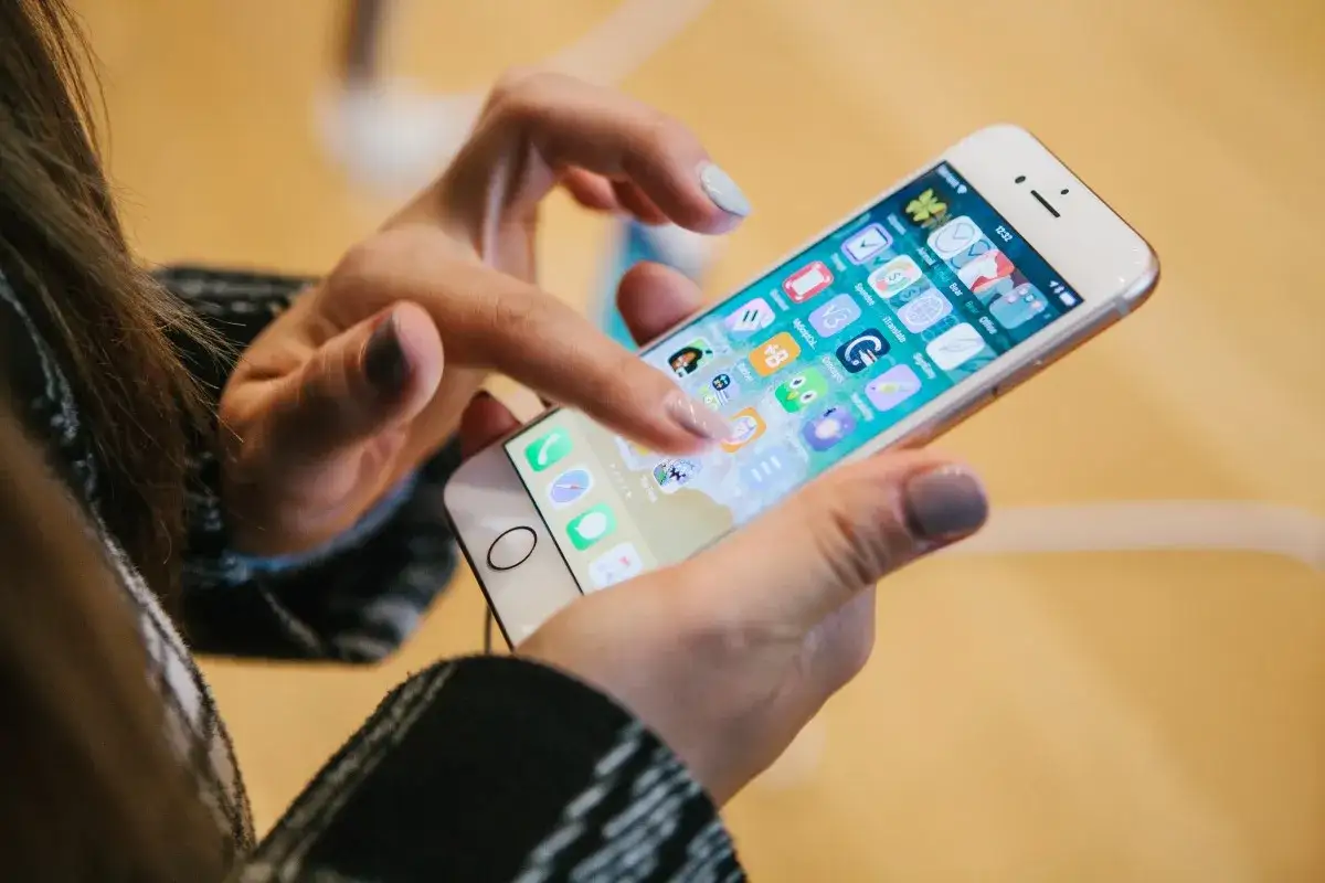 Dłoń z paznokciami pomalowanymi na jasny i ciemny szary kolor dotyka ekranu telefonu z ikonami aplikacji, widoczny jest interfejs systemu iOS 15.